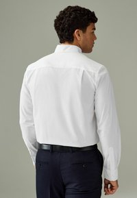 Chemise blanche à manches longues avec un col standard et une ceinture noire, portée par-dessus un pantalon foncé, mettant en valeur une texture lisse avec un design ajusté.