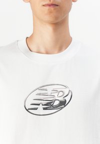 Balta medvilninė marškinėlių su dideliu, blizgančiu sidabriniu ovaliu logotipu, kuriame yra stilizuota "NB" rašyba ir žvaigždžių dizainas priekyje.