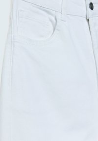 Gros plan d’un pantalon en denim blanc montrant la poche avant, la petite poche à monnaie, la boucle de ceinture et la fermeture par bouton métallique