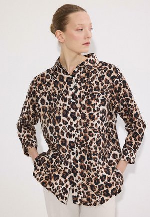 Femme aux cheveux attachés portant une chemise à manches longues à imprimé léopard, les mains dans les poches, regardant sur le côté.