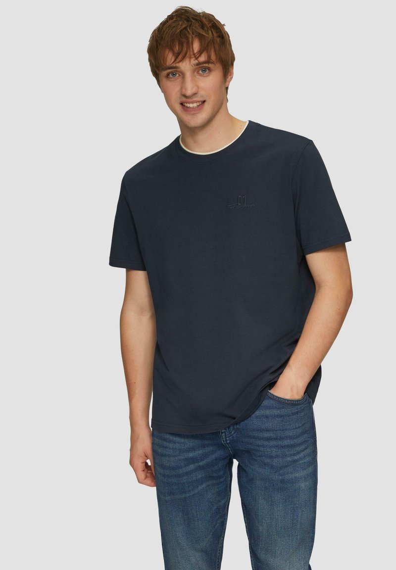T-shirt en coton bleu marine avec un col rond et des manches courtes, présentant un logo brodé subtil sur la poitrine. Porté avec un jean bleu.
