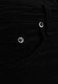 Jeans noirs avec une texture lisse, design à cinq poches, matériel argenté et détails de couture subtils. Accent sur la taille et la zone du bouton.