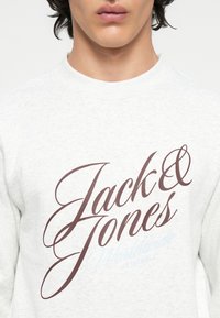 Világosszürke pulóver, amelyen a "Jack & Jones" sötétbarna írással és kék díszítéssel látható. Puha anyag, bordás nyakrésszel.