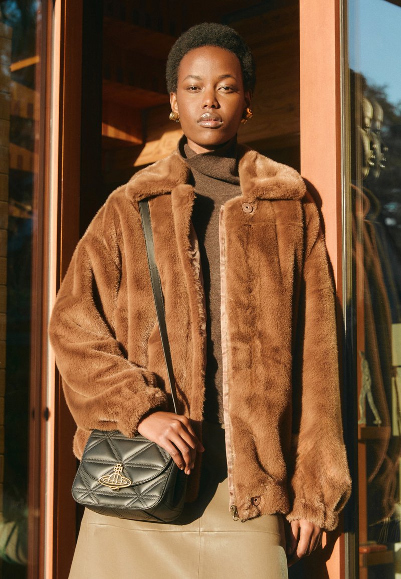 Uma jaqueta de pele sintética castanha com colarinho, fecho de correr e punhos ribados. Uma mala de ombro preta acolchoada com um detalhe de logótipo. Modelo vestindo uma blusa castanha.