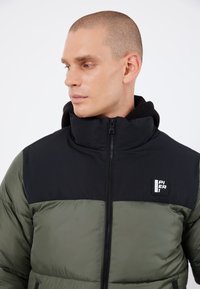 Chaqueta acolchada con una sección superior negra y una inferior de color verde oliva. Cierre de cremallera, cuello alto y parche con logo en el pecho. Textura suave.