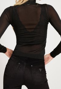 Un col roulé noir transparent à manches longues présente un accent en dentelle au dos. Associé à un pantalon en velours côtelé noir avec des textures subtiles et des poches.