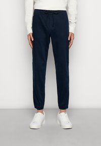 Marinblå joggerbyxor med elastiska muddar, dragsko i midjan och sidofickor, tillverkade av mjukt, lätt bomullstyg. Vita sneakers.