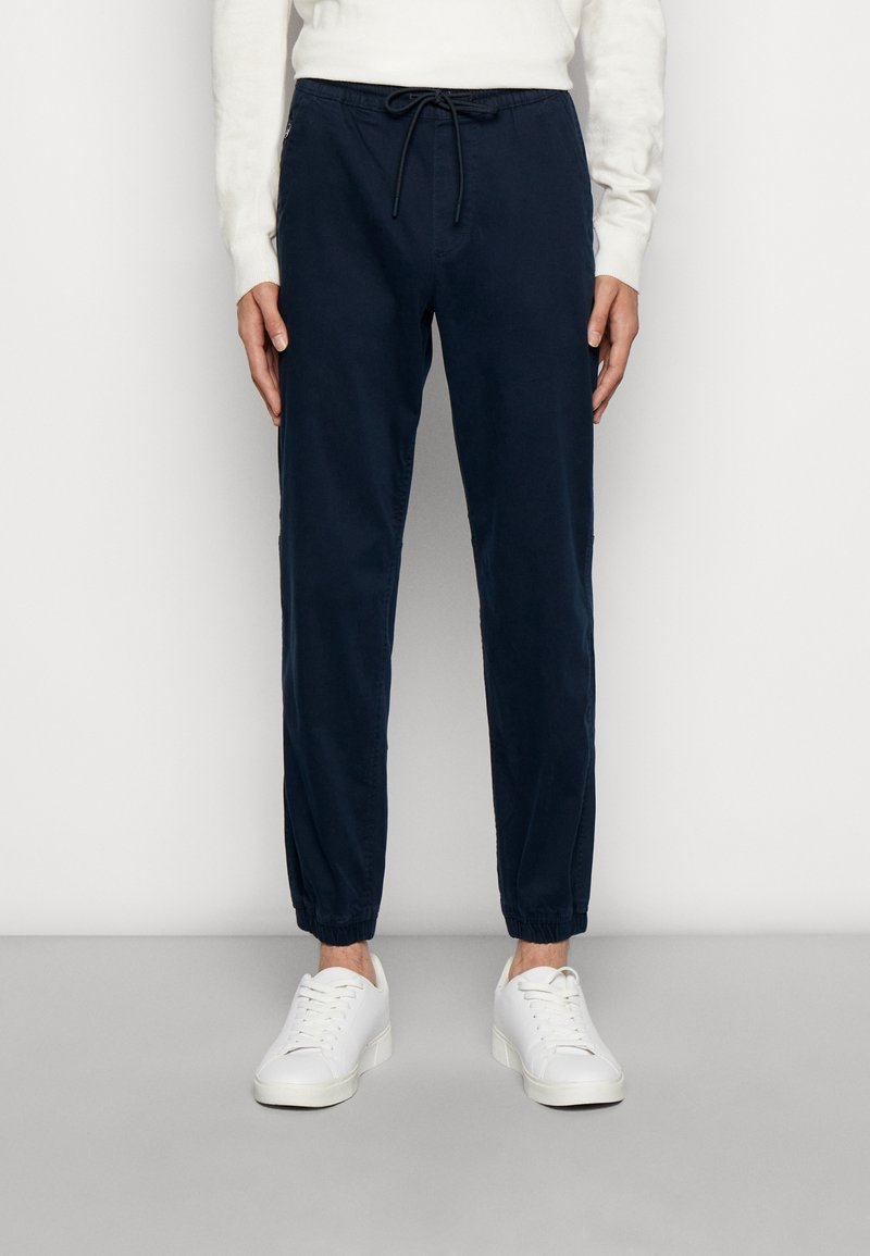 Marinblå joggerbyxor med elastiska muddar, dragsko i midjan och sidofickor, tillverkade av mjukt, lätt bomullstyg. Vita sneakers.