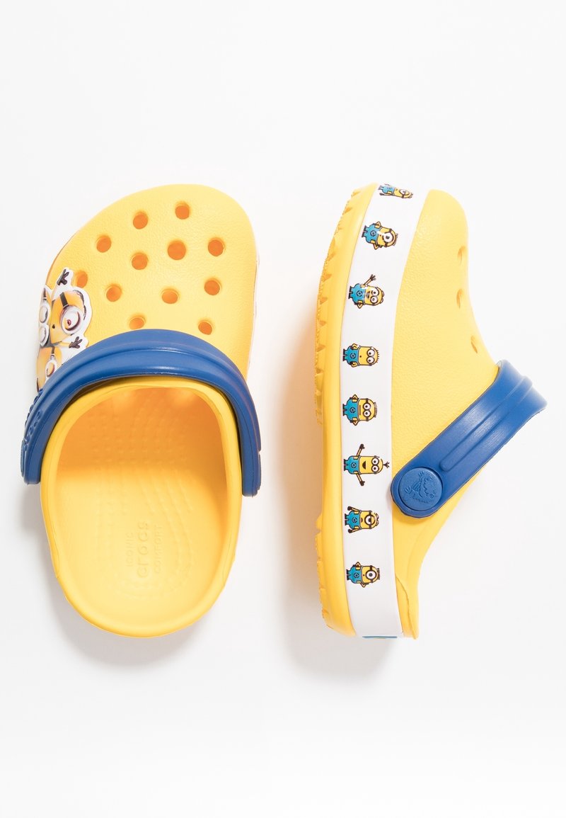 Gula träskor gjorda av syntetiskt material med ventileringshål, blå rem och Minion-karaktärer på sidorna.