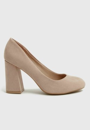 Zapato de tacón alto de ante color nude, con puntera redondeada, tacón grueso y textura suave. Presenta un diseño sencillo y líneas limpias.