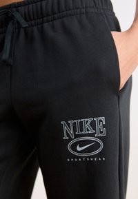 Melni Nike sporta bikses ar elastīgu jostu, sānu kabatām un druku ar logotipu, kurā ir uzraksti "NIKE" un "SPORTSWEAR" baltā krāsā.
