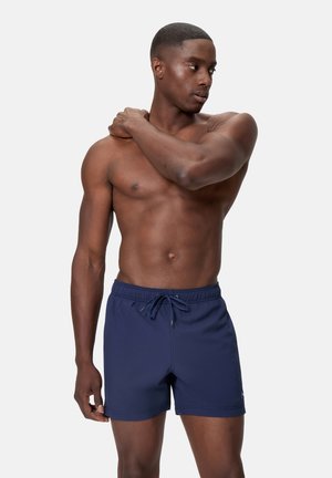 Uomo muscoloso con pantaloncini da bagno blu navy, in piedi a torso nudo, che tiene la spalla sinistra con la mano destra mentre guarda a destra.