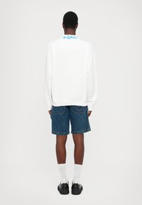 Λευκό oversized sweatshirt με την ένδειξη "MILANO" τυπωμένη σε μπλε στην πλάτη, συνδυασμένο με τζιν σορτς. Το μοντέλο φορά μαύρα παπούτσια και λευκές κάλτσες.