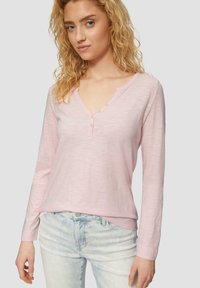 Lichtroze langarm Henley shirt met een V-hals en drie knopen bij de kraag. De stof heeft een zachte textuur; gecombineerd met lichtblauwe jeans.
