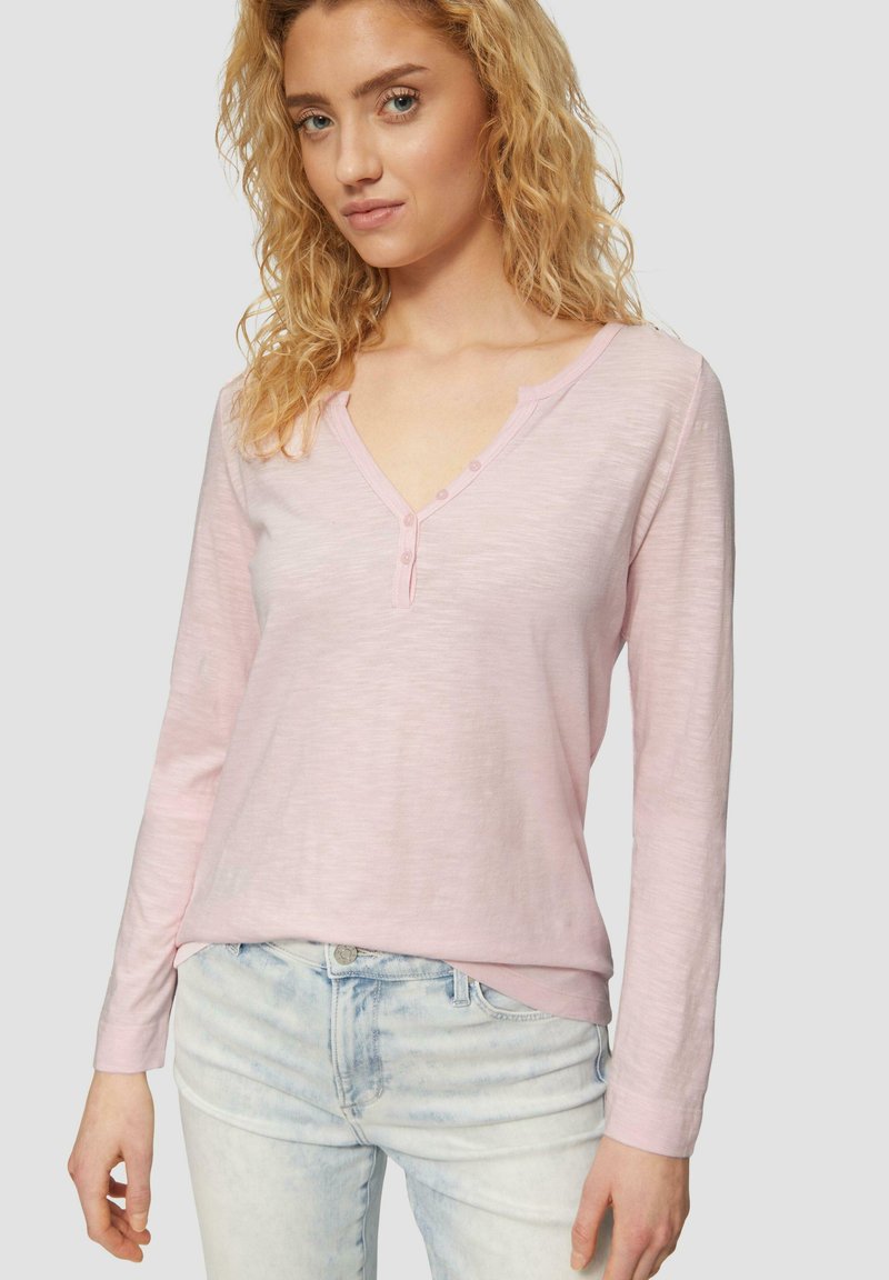 Lichtroze langarm Henley shirt met een V-hals en drie knopen bij de kraag. De stof heeft een zachte textuur; gecombineerd met lichtblauwe jeans.