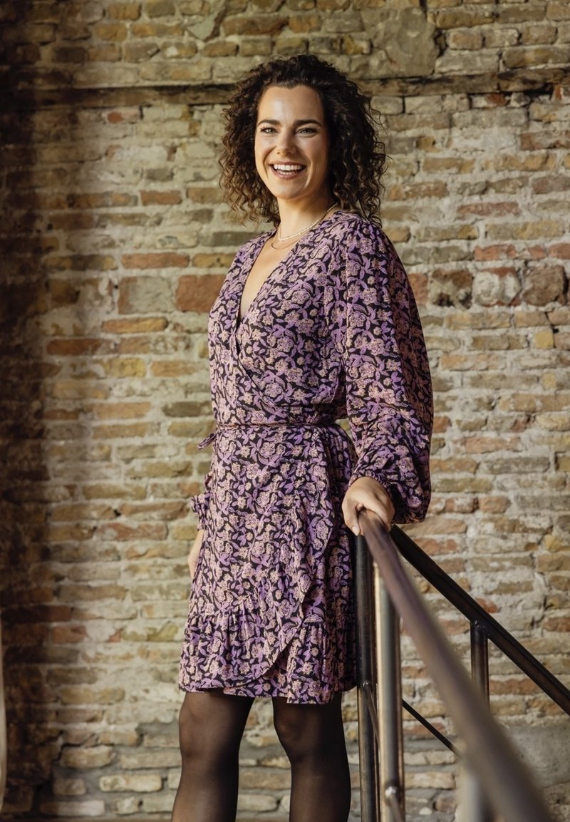 Paarse wikkeljurk met bloemmotieven, ruches aan de onderkant en lange mouwen. Gekozen met zwarte panty’s, staand tegen een stenen muur.