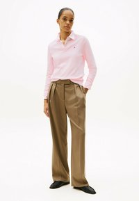 Rosa Langarm-Polo mit V-Ausschnitt und Logo, kombiniert mit hoch taillierten, braunen Plisseehosen. Schwarze Schuhe runden das Outfit ab.