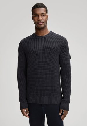 Junger Mann, der frontseitig steht, trägt einen dunkel strukturierten Strickpullover und dunkle Hosen vor einem schlichten Hintergrund.