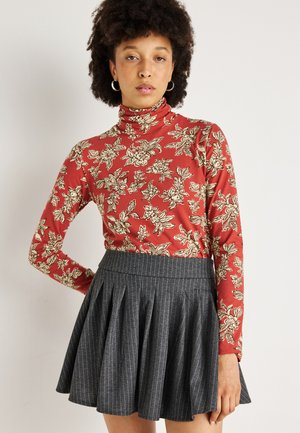 Raudona gėlių modelio golfas turi ilgomis rankovėmis ir prigludusią siluetą, derinamas su pilku dryžuotu klostuotu sijonu. Minkšta audinio tekstūra.