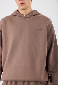 Brązowa bluza z kapturem wykonana z miękkiego materiału, z logo na przodzie w dopasowanym odcieniu, luźnym krojem oraz regulowanym kapturem ze sznurkiem.