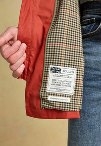 Raudonas išorinis audinys, sijonas su patikrinta vidine pamušalu. Yra etiketė su Britų vėliava ir priežiūros instrukcijomis. Matomi tekstūruoti medžiagų paviršiai.