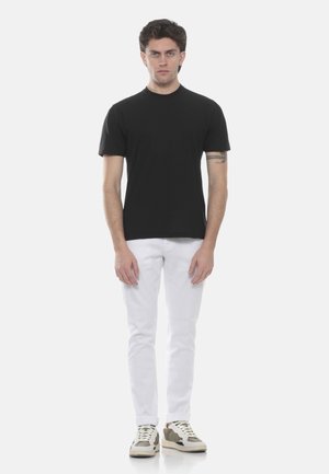 Giovane uomo in piedi, indossa una maglietta nera, pantaloni bianchi e sneakers beige con lacci, su uno sfondo chiaro uniforme.