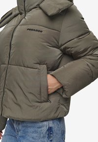 Nafukovacia olivovozelená bunda s vysokým golierom, zipsom a viditeľným šitím. Má malé logo a bočné vrecká.