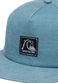 Blå denimkeps med en svart och vit broderad logotypapplikation med vågmönster. Platt skärm och texturerad tygdetalj.