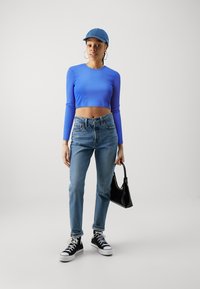 Un crop top bleu à manches longues, un jean bleu clair taille haute, des baskets montantes noires et un sac à main noir, sur un fond uni.