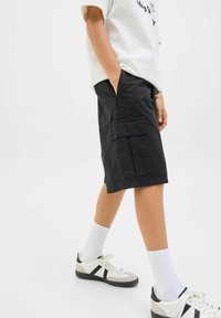 Shorts cargo noirs avec poches latérales, fabriqués à partir d'un tissu résistant. Portés avec un t-shirt blanc et des baskets sportives, mettant en avant une coupe décontractée.
