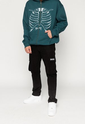 Personne portant un sweat à capuche zippé turquoise avec un graphique blanc de cage thoracique et un pantalon cargo noir avec un patch "Multiply Apparel", debout, main dans la poche.