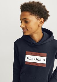 Sudadera marina con un bolsillo tipo canguro, que presenta un logo cuadrado en rojo y blanco que dice "JACK & JONES" y "ORIGINALS WORLDWIDE."