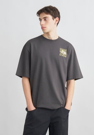 Giovane uomo con indosso una maglietta oversize grigio scuro con una grafica gialla sul petto, in piedi con le mani nelle tasche, che guarda di lato.