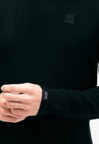 Schwarzer Pullover aus weichem Strickmaterial, mit rundem Halsausschnitt und enganliegenden Bündchen, versehen mit einem Markenlabel am Ärmel.