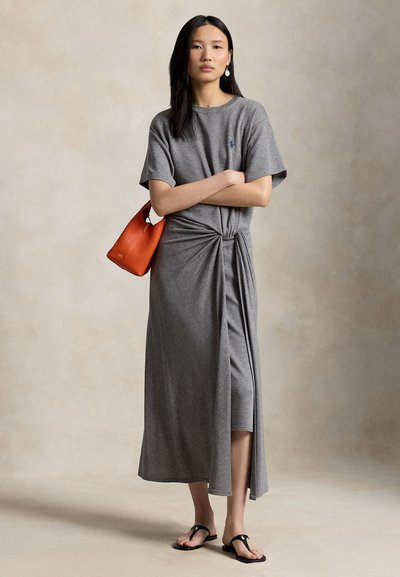 Mujer con cabello largo y oscuro, vestida con un vestido midi gris anudado, sandalias negras, pendientes blancos colgantes y sosteniendo un bolso naranja en el brazo.