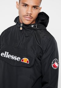 Svart, lett jakke med halv glidelås-design, en frontlomme og broderte logoer. Har hette og en glatt, shiny tekstur.