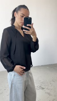 Černá poloprůsvitná blůza s výstřihem do V a elastickými manžetami, kombinovaná s volnými světlomodrými džíny, které zdůrazňují uvolněnou siluetu.