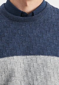 Pletený svetr s texturovaným vzorem v tmavě modré a světle šedé barvě, s kulatým výstřihem a žebrovanými manžetami. Pod ním je tmavě modrá košile s puntíky.