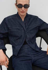 Veste en denim bleu foncé avec un col, deux poches avant et fermeture à boutons. Portée sur un haut noir, assortie avec un pantalon en denim foncé.