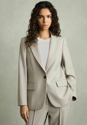 Beige maßgeschneiderter Blazer mit Reverskragen, einer Knopfleiste, zwei vorderen Taschen und einer glatten Textur, kombiniert mit einem weißen T-Shirt.