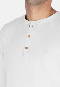 Weißes Langarmhemd mit runder Kragen, drei Holzknöpfen und einem gestickten Logo auf der linken Seite. Glatte Baumwolltextur.