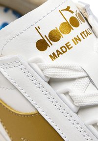 Sneaker bianco con dettagli dorati, con tomaia in pelle testurizzata, lacci bianchi piatti e logo stampato che recita "Fatto in Italia".