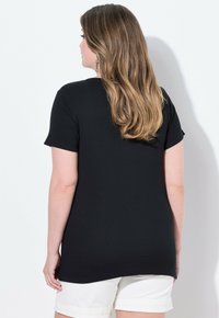 T-shirt nera a coste con maniche corte, orlo arrotondato e vestibilità casual, mostrata da dietro abbinata a pantaloni corti di colore chiaro.