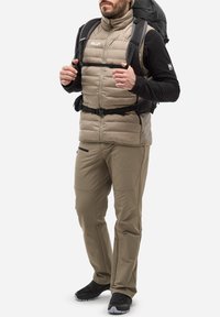 Beige isolierte Weste über einem schwarzen Langarmshirt, kombiniert mit hellbraunen Hosen und schwarzen Schuhen. Ausgestattet mit einem schwarzen Rucksack und Riemen.