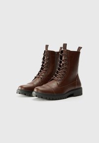 Botas de cuero marrón hasta el tobillo con cordones en la parte delantera, punta redondeada, costuras pronunciadas y un acabado texturizado; cuentan con una suela de goma con relieves.