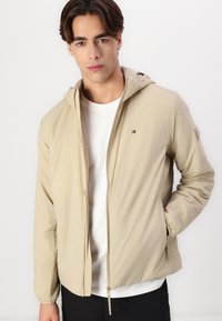 Lys beige hettejakke med glidelås, laget av et glatt materiale. Har elastiske mansjetter og en liten logo på brystet. Brukes over en hvit skjorte.