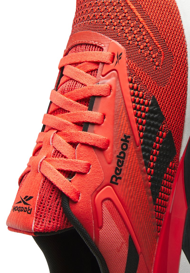 Zapatilla atlética roja con parte superior de malla texturizada, acentos negros y una destacada marca Reebok. Presenta cordones brillantes y una suela acolchada.