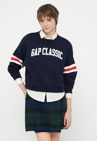 Sudadera de color Navy con "GAP CLASSIC" en texto blanco, con franjas rojas y blancas en las mangas. Combinada con una falda de cuadros en verde y azul.