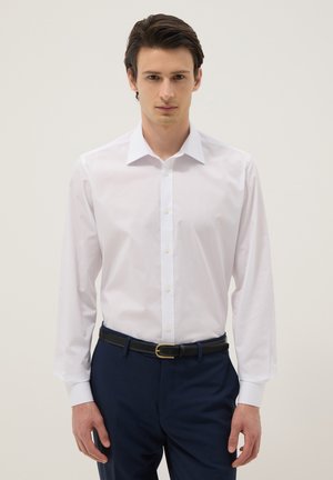 Junger Mann trägt ein weißes Hemd, das in marineblaue Hose mit schwarzem Gürtel gesteckt ist, steht vor einem schlichten hellen Hintergrund.