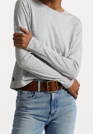 Persoon draagt een lichtgrijs shirt met lange mouwen met een klein logo, blauwe spijkerbroek en een bruine leren riem met een vierkante gouden gesp.